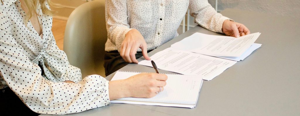 Two Women Sitting at Grey Table Top Writing on Notebook and Reviewing Tax Documents