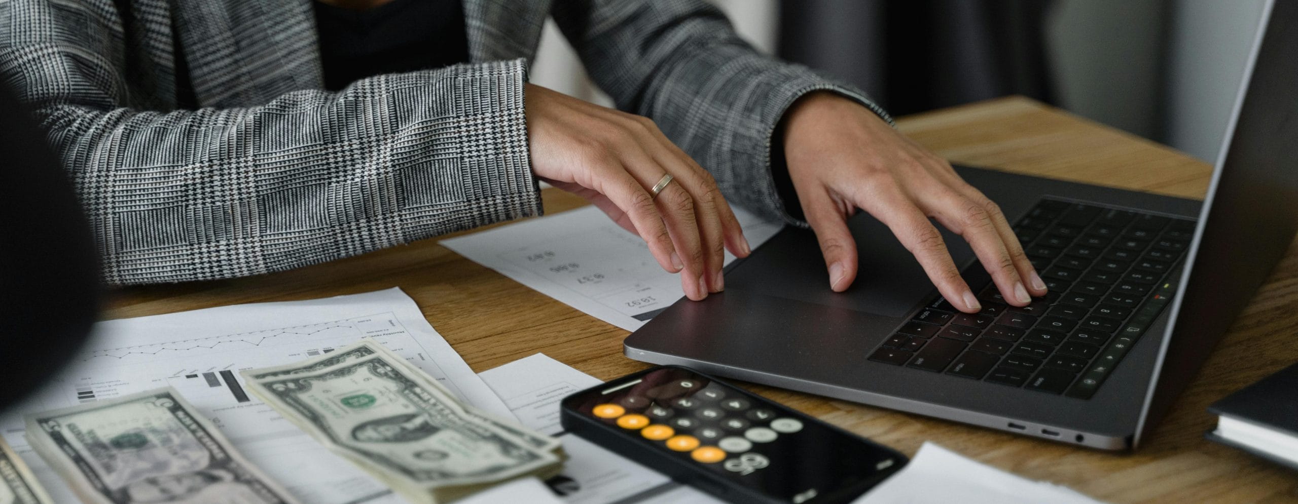 woman typing on laptop computer with calculator, cash and financial documents sitting on dark wooden surface.