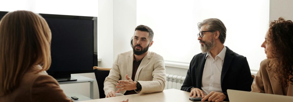 Individuals sitting around a conference table in an office dressed in business attire with the window light shining in on them.