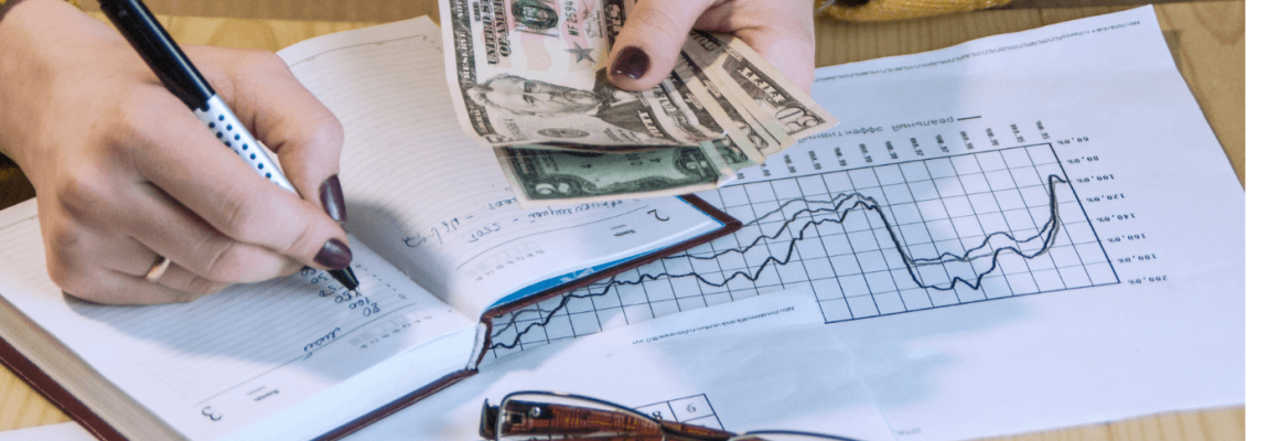 Money in hand of a younger lady writing on notebook and financial documents around on table top.