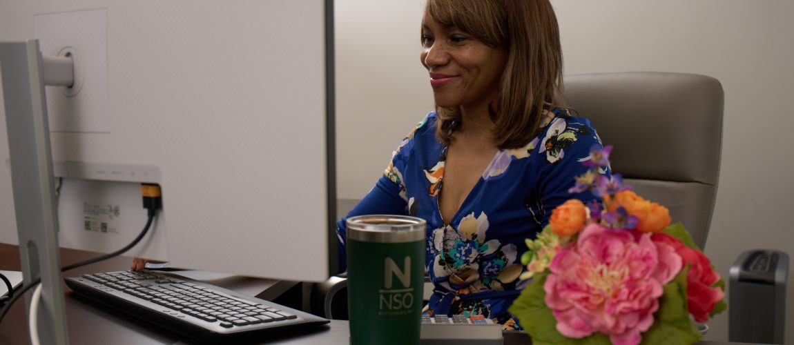 Nina, CPA, sitting at her dark wood desk, looking at her desktop monitor with flowers and a branded mug on her desk.