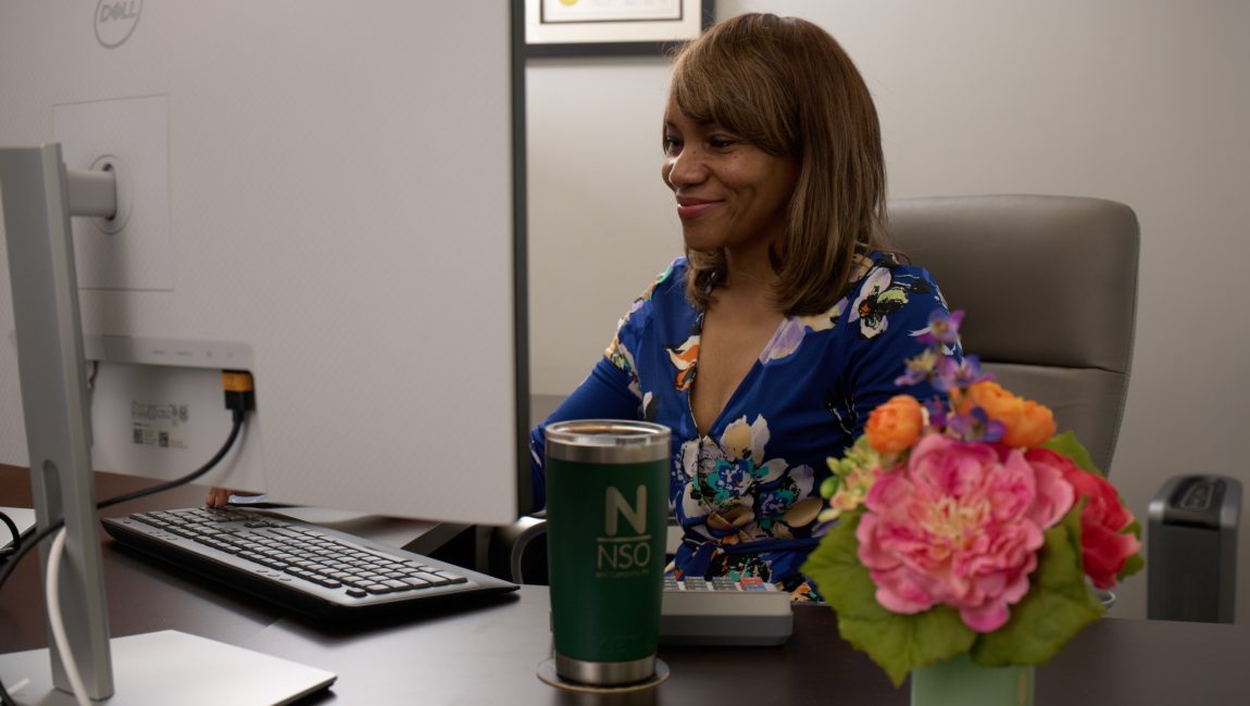 Nina, CPA, sitting at her dark wood desk, looking at her desktop monitor with flowers and a branded mug on her desk.