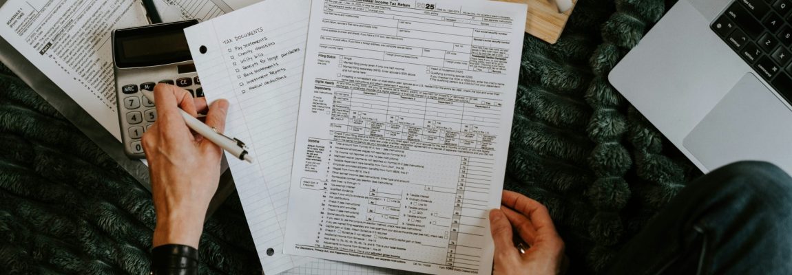 Hand holding tax documents in front of dark fuzzy blanket with computer and office supplies nearby