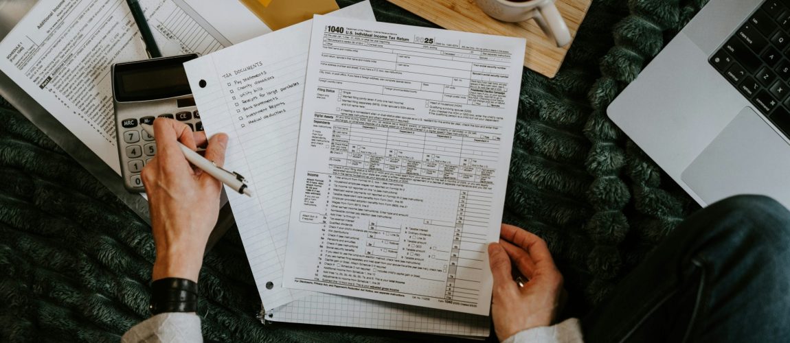 Hand holding tax documents in front of dark fuzzy blanket with computer and office supplies nearby