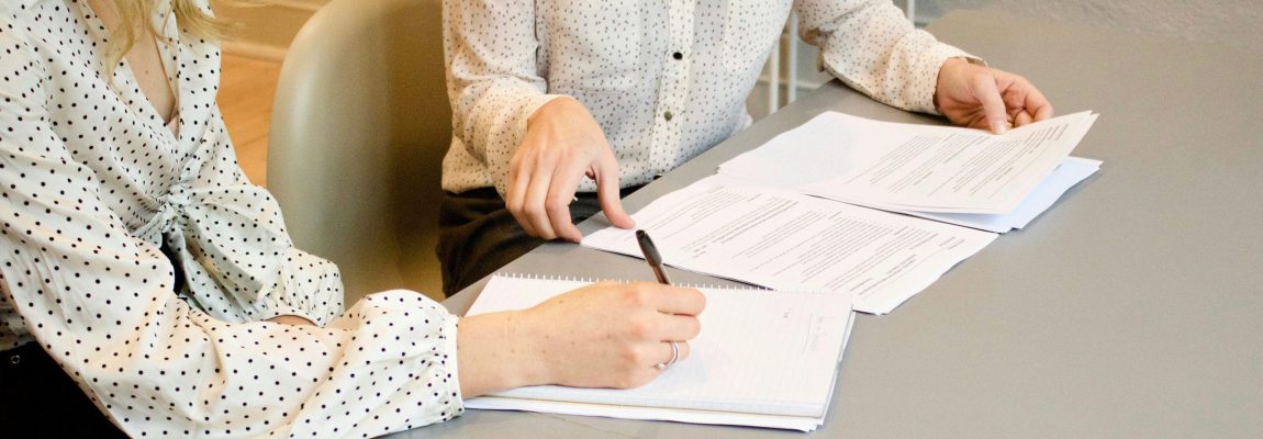 Two Women Sitting at Grey Table Top Writing on Notebook and Reviewing Tax Documents