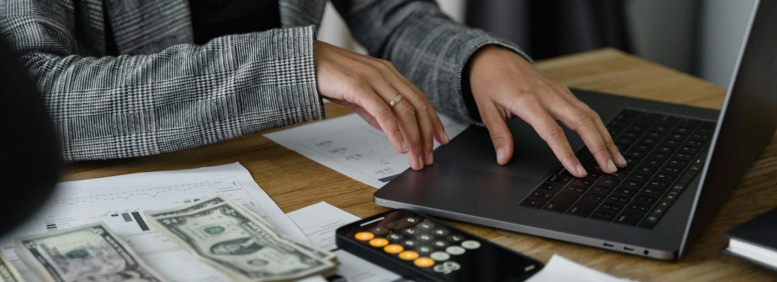woman typing on laptop computer with calculator, cash and financial documents sitting on dark wooden surface.
