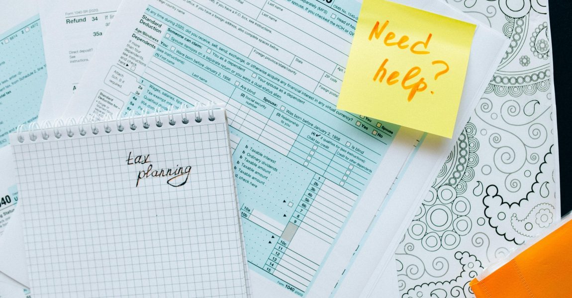 white and light blue tax documents layed out on a table top with a yellow sticky note and red pen writing.